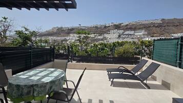 Un patio al aire libre con una mesa y sillas, vista a una colina con edificios y una cerca verde.
