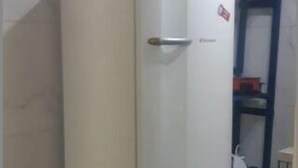Geladeira compacta e branca num canto de um quarto com chão em azulejos e parede branca.