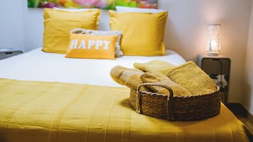 Una cama bien hecha con ropa de cama y almohadas amarillas, una cesta con toallas dobladas y una lámpara fijada a la pared.