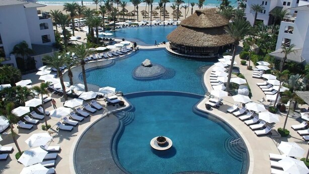 Ein Resort-Pool mit Liegestühlen und Sonnenschirmen, umgeben von Palmen und einem Strand.