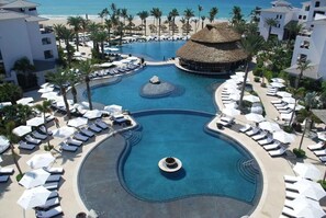 Piscina de resort con sillones de descanso y sombrillas, rodeada de palmeras y una playa.