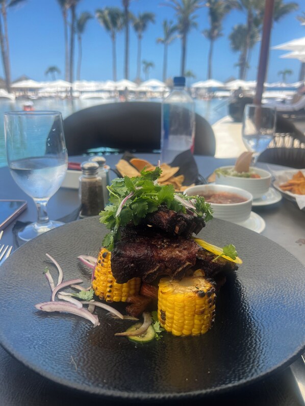 Una mesa de comedor con un plato de carne a la parrilla y maíz, rodeada de diferentes salsas y un vaso de agua.