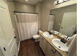 A master bathroom with a toilet, two sinks, a shower with a curtain, and a mirror above the sink.