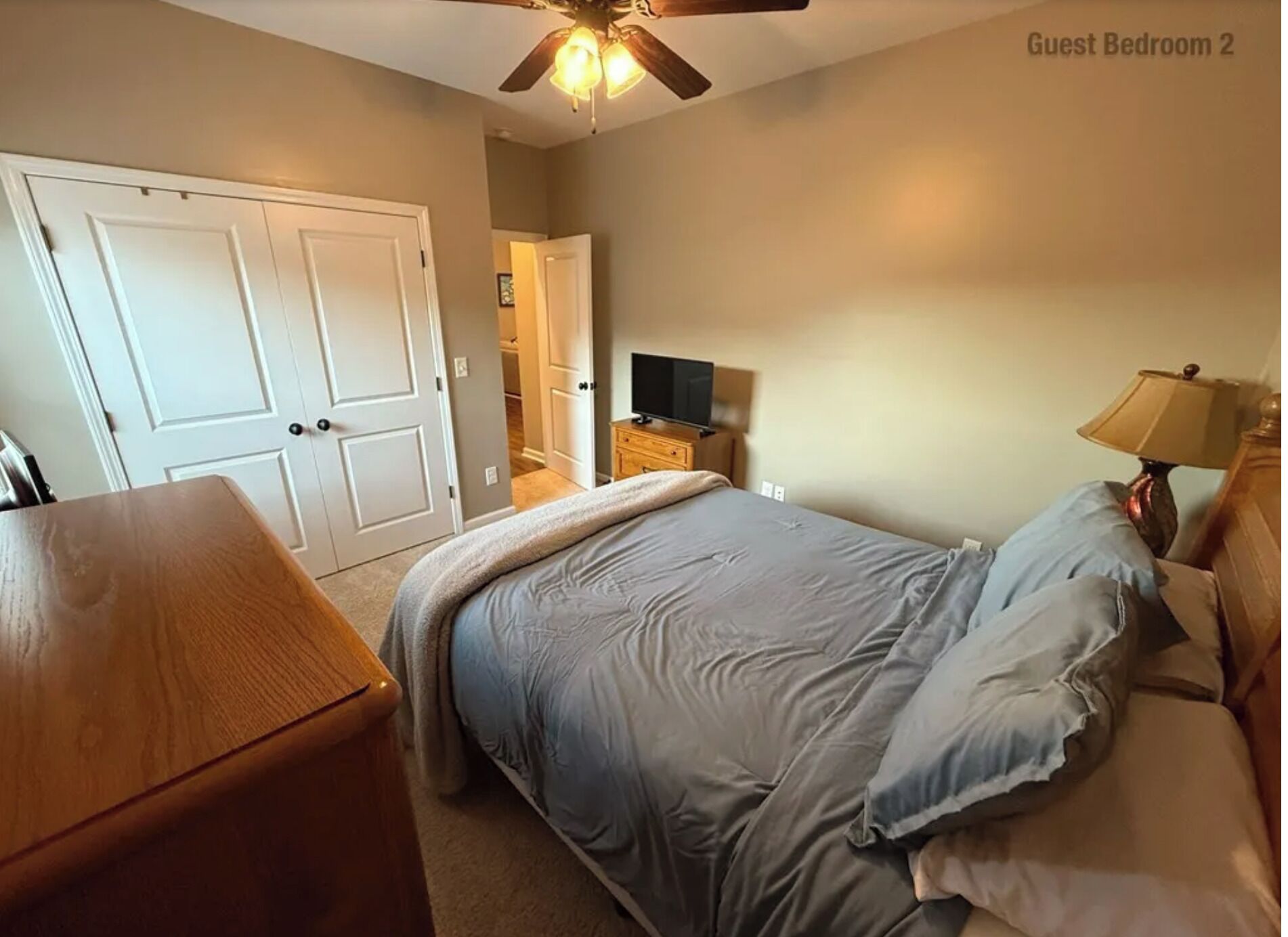 Une chambre d’amis comprenant un lit, une table de chevet en bois, un téléviseur et un ventilateur de plafond.