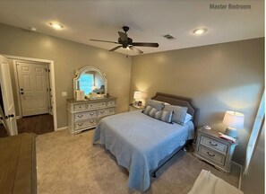A master bedroom with a large bed, a dresser, a vanity with a mirror, a ceiling fan, and a nightstand with a lamp.