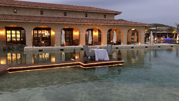 Un espace aménagé au bord de la piscine avec des tables et des chaises, un bâtiment doté de fenêtres en arc de cercle et une piscine à l’eau claire.