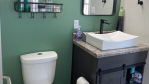 Une salle de bain équipée de toilettes, d’un lavabo, d’un miroir et d’une étagère où sont rangés les articles de toilette.