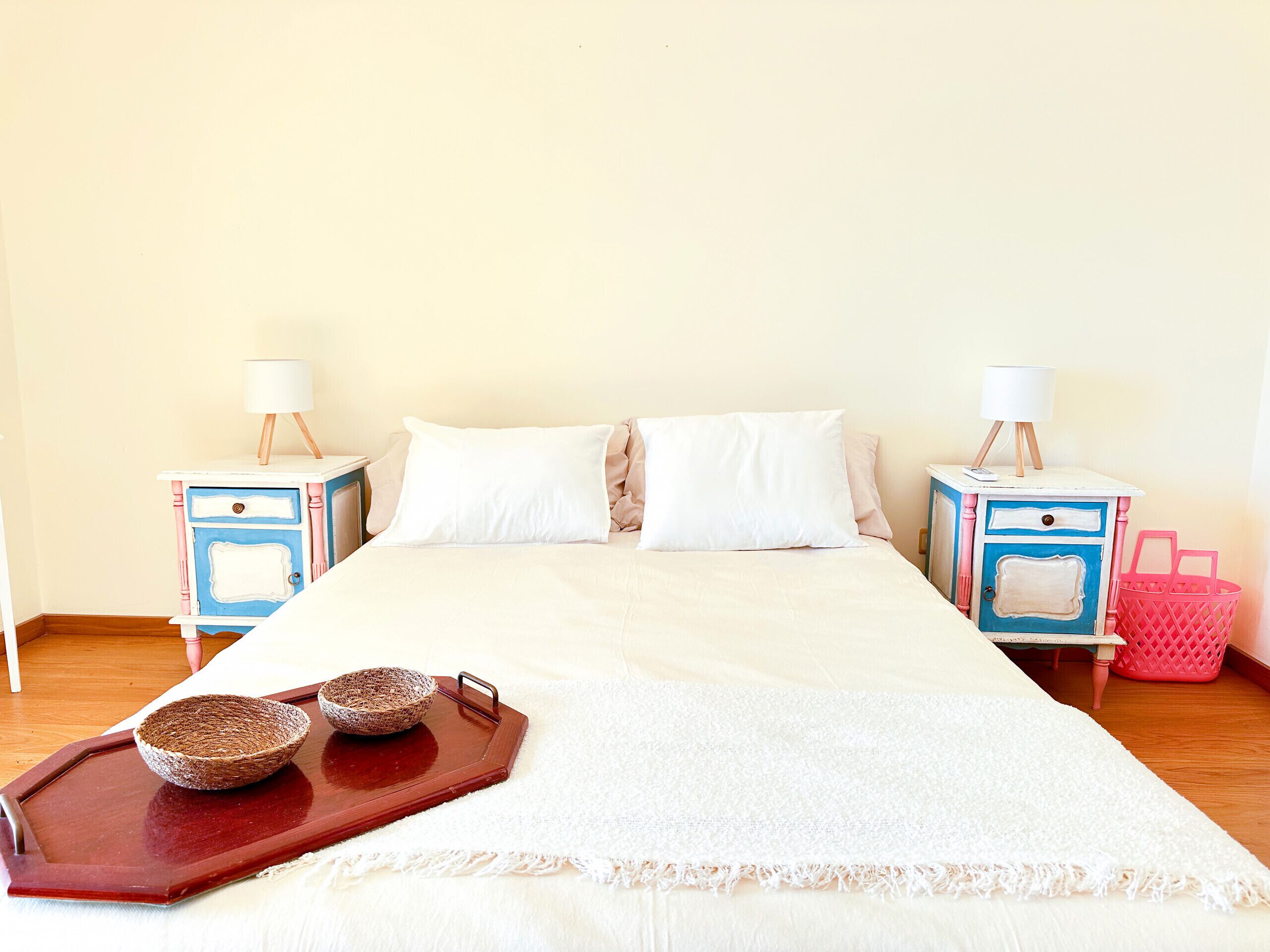 Cama bem arrumada com lençóis brancos, duas mesas de cabeceira com abajures e um cesto de madeira com duas tigelas no chão.