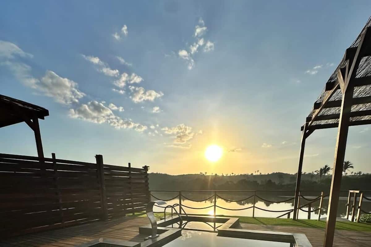 Um deck de madeira com hidromassagem e cerca de madeira, com vista para um corpo d'água e um pôr do sol.