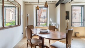 Un espace repas avec une table et des chaises en bois, de grandes fenêtres offrant une vue sur les bâtiments et des lampes suspendues.