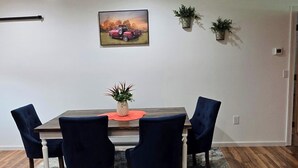 Uma área de jantar com uma mesa de madeira, quatro cadeiras azul-escuras, um jogo americano vermelho e uma planta em vaso sobre a mesa.