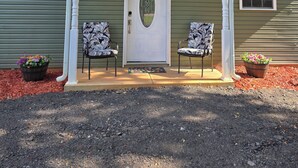 Uma casa com fachada verde, uma porta da frente branca com painel de vidro decorativo, duas cadeiras com almofadas estampadas e uma pequena janela à direita da porta.