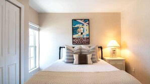 Cama bem arrumada com almofadas listradas, um cartaz com cena de praia e abajur.