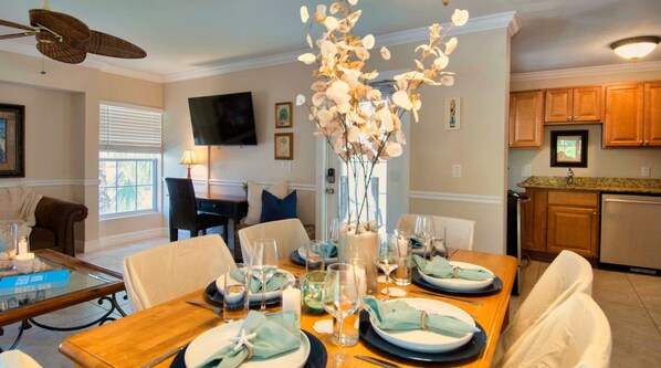 A dining area with a wooden table set for four, a vase with white flowers, and a television mounted on the wall.