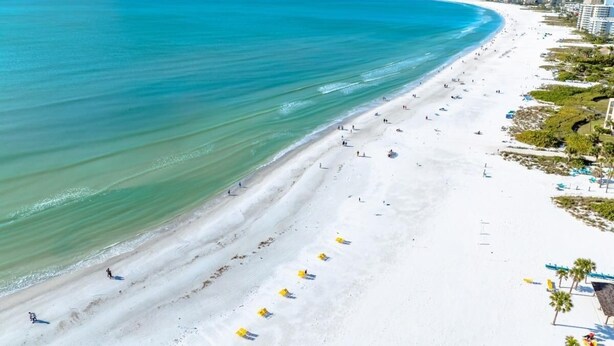 Uma praia extensa com águas azul-claras, areal arenoso e alguns edifícios ao fundo.