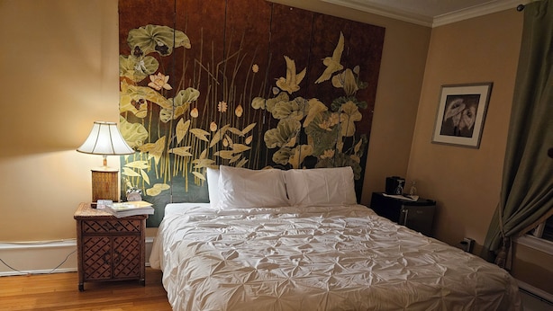 A neatly made bed with a white duvet, a bedside table with a lamp, and a wall with a floral tapestry. - TTR - Kensington (Pembroke)