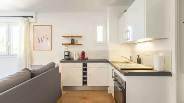 Een compacte keuken met witte kastjes, een houten aanrecht en een inbouwoven. In de aangrenzende woonkamer staat een grijze bank.