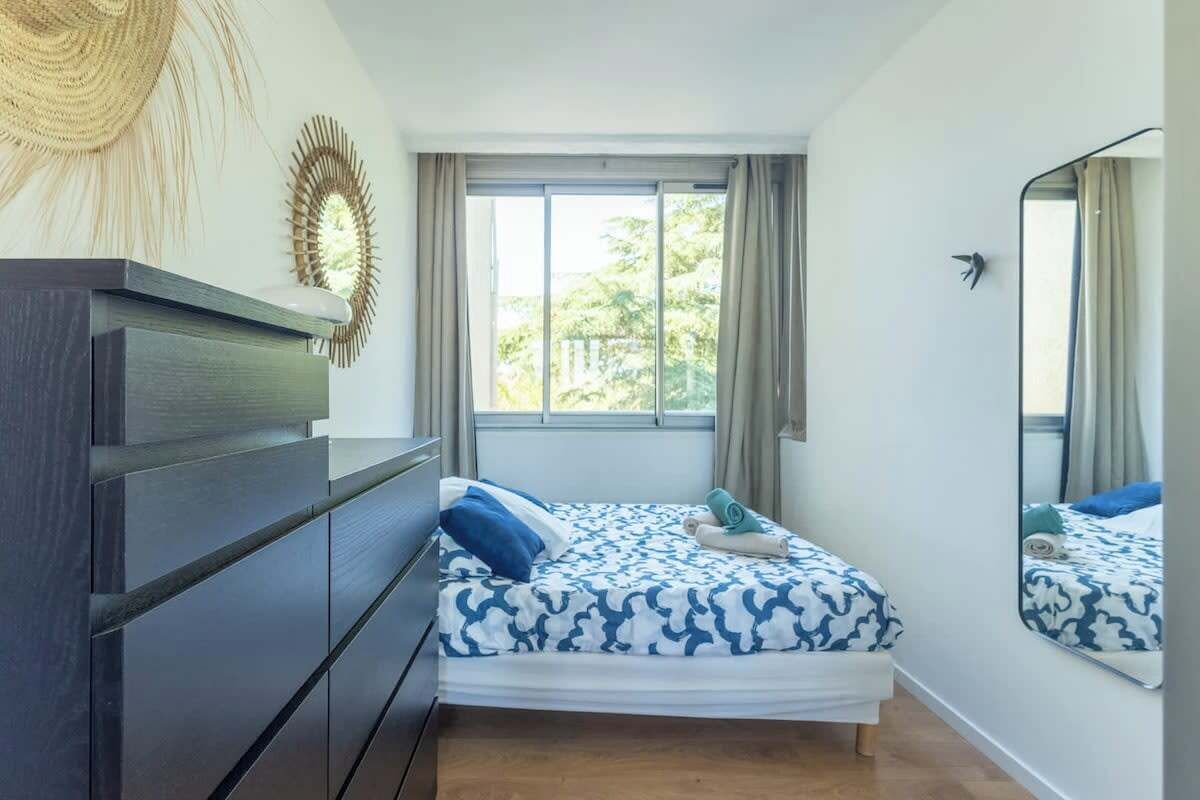 Ein Schlafzimmer mit Bett, Kommode, einem Fenster mit Vorhängen und einem Spiegel.