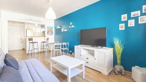 A modern living room with a blue accent wall, a flat-screen TV, a white entertainment unit, and a dining area with white chairs and a table. - Nid du 5ème - Proche Bd Baille - Métro et commerce (Marseille)