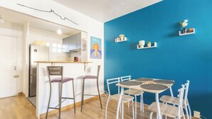 A compact kitchen with a wooden countertop and two bar stools, a dining table with four chairs, and a blue accent wall with shelves holding decorative items. - Nid du 5ème - Proche Bd Baille - Métro et commerce (Marseille)