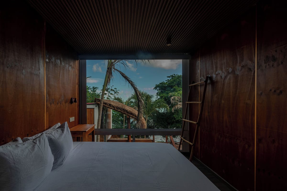 A room with a bed, wooden walls, a window with a view of palm trees and a thatched-roof structure.