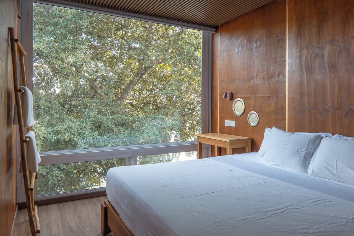 A bedroom with a large window, wooden walls, a bed with white linens, a small wooden table, and a wall-mounted clock.
