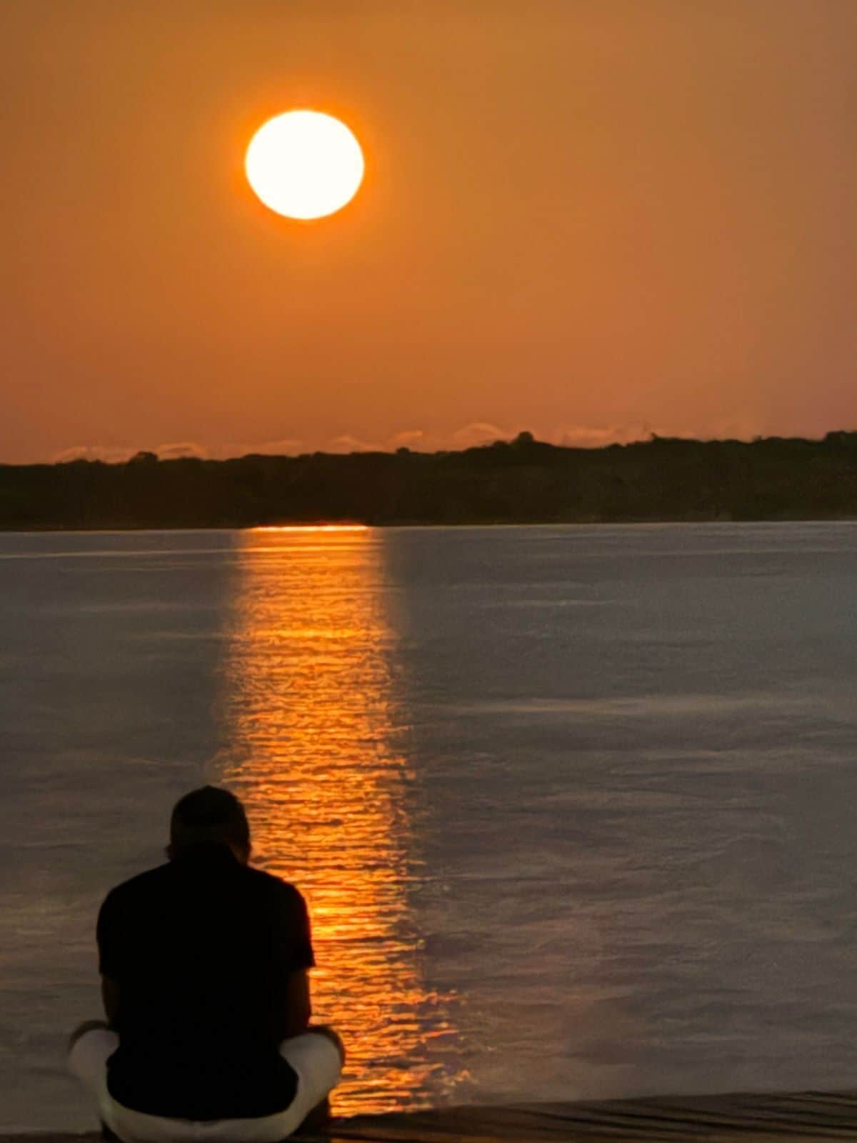 A person sitting by a calm body of water during sunset, with the sun reflecting off the water's surface.