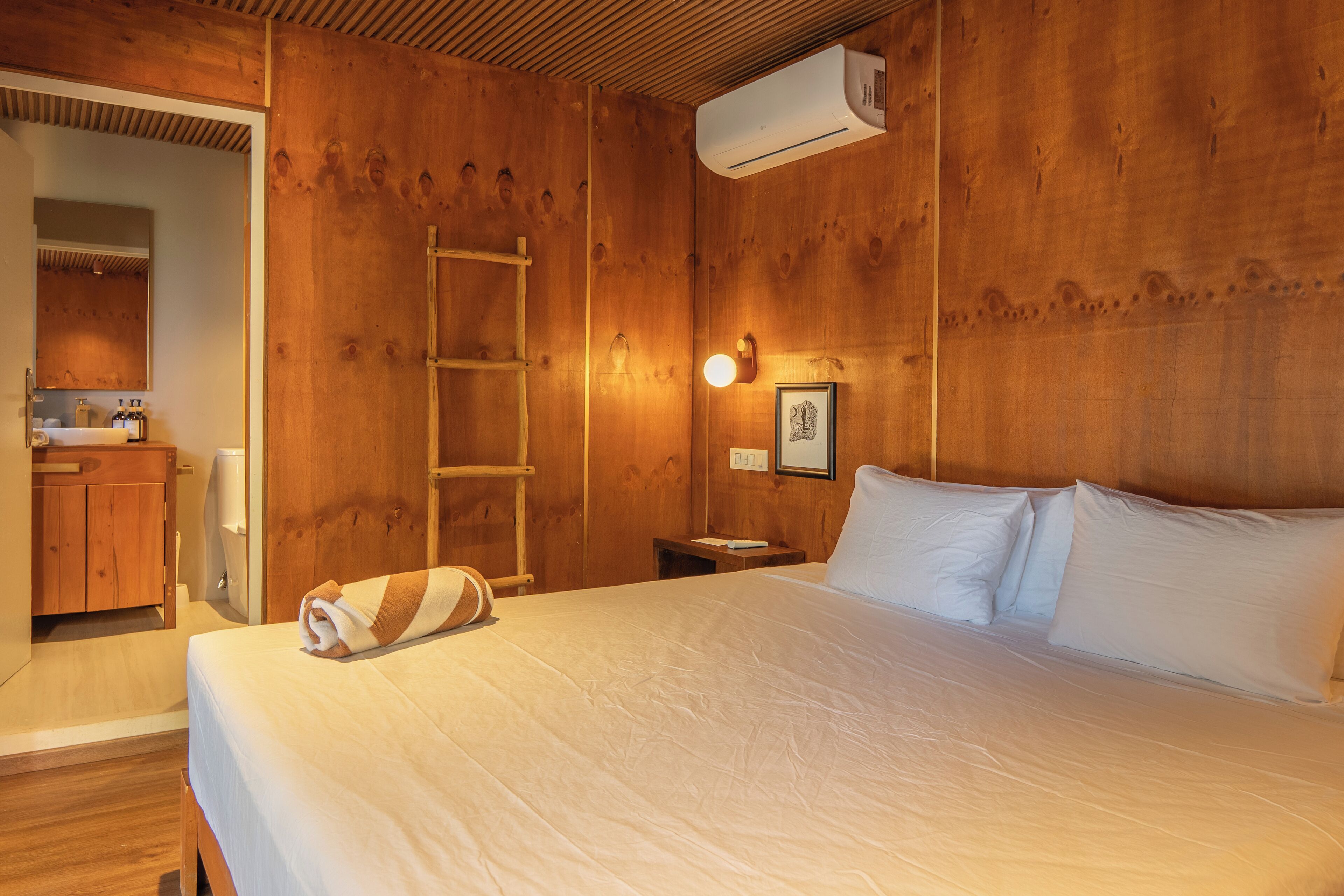 Quarto com painéis de madeira, uma cama, mesa de cabeceira, ar condicionado fixado na parede, quadro emoldurado e um armário de madeira com espelho.