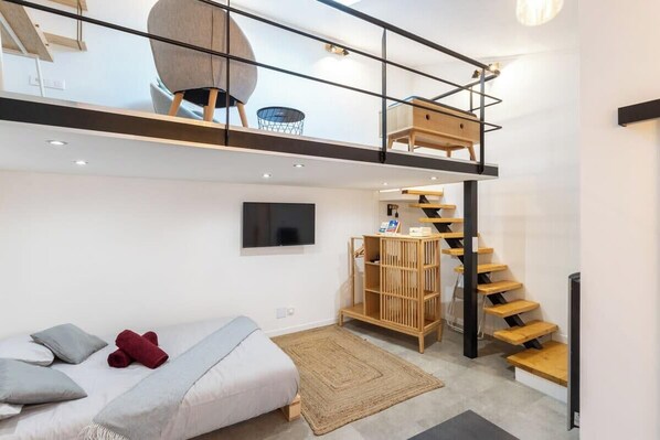 A modern loft bedroom with a loft bed, a built-in TV, a wooden staircase, and a small seating area with a chair and table.