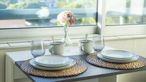 Uma mesa posta com pratos, copos e um vaso de flores, posicionada junto a uma janela com vista para o verde.