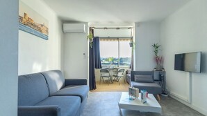 Uma sala de estar moderna com um sofá cinzento, uma mesa de centro e uma televisão na parede. Há uma varanda com mesa e cadeiras, e um quadro com uma paisagem costeira na parede.
