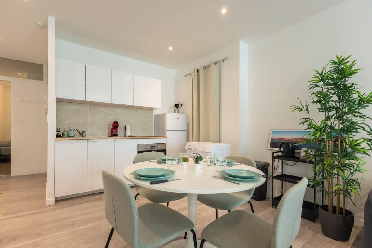 Une cuisine moderne avec des meubles blancs, une table à manger dressée avec de l’assiette turquoise et une plante en pot.