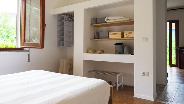 Ein Schlafzimmer mit Bett, eingebauten Regalen mit Aufbewahrungsboxen, einem Wäscheständer und einem Fenster mit Holzrahmen.