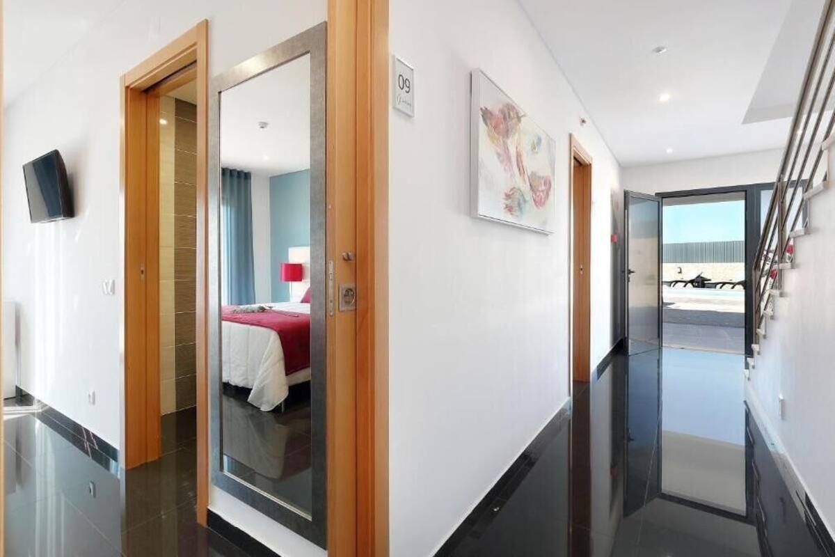 Un couloir d’hôtel avec un mur recouvert de miroirs, qui reflète une chambre avec un lit et une couverture rouge. Un tableau est accroché au mur, et un téléviseur y est fixé.