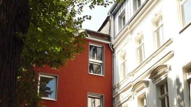 A narrow street with a red building and a white building with columns. - Apartment in In-Veedel Ehrenfeld (Köln)
