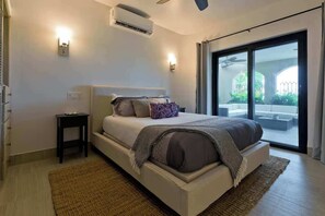 Une chambre moderne avec un lit, des tables de chevet, un appareil de climatisation et une vue dégagée sur l’extérieur grâce à une grande porte-fenêtre coulissante.