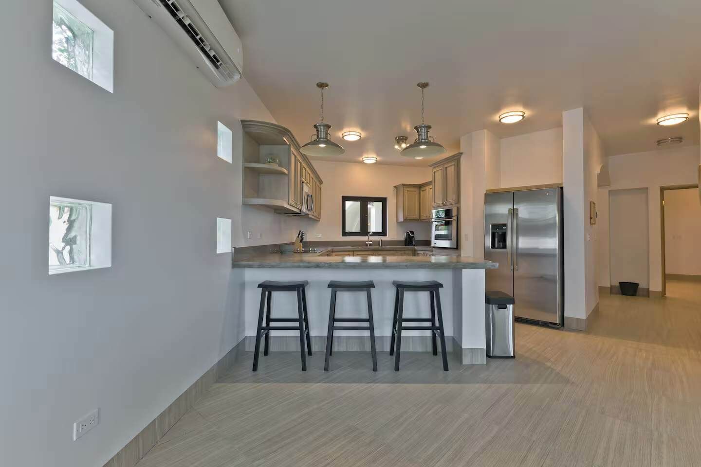 A modern kitchen with a central island, bar stools, built-in cabinets, a stainless steel refrigerator, and pendant lighting.