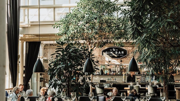 A restaurant interior with tables set for dining, a bar area, and large indoor plants.