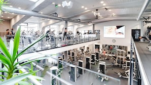 A multi-level gym with various exercise equipment, a large screen, and a glass railing overlooking the lower level.