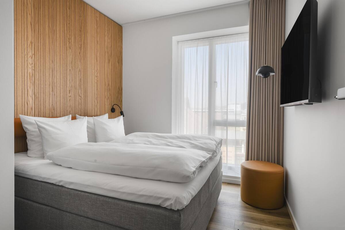 A modern bedroom with a bed, a round ottoman, a TV mounted on the wall, and a large window.