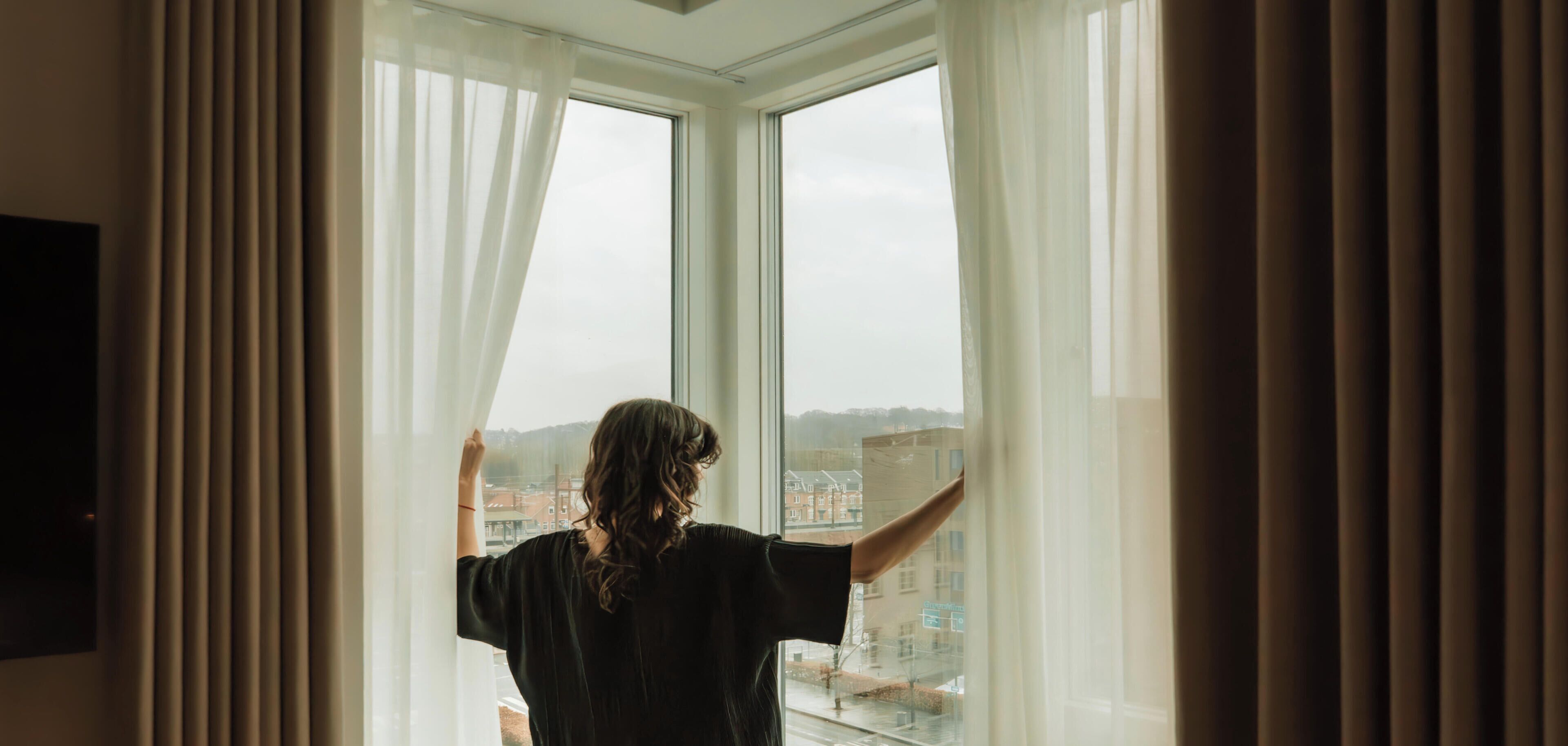 A person standing by a window with sheer curtains, looking out at a cityscape.