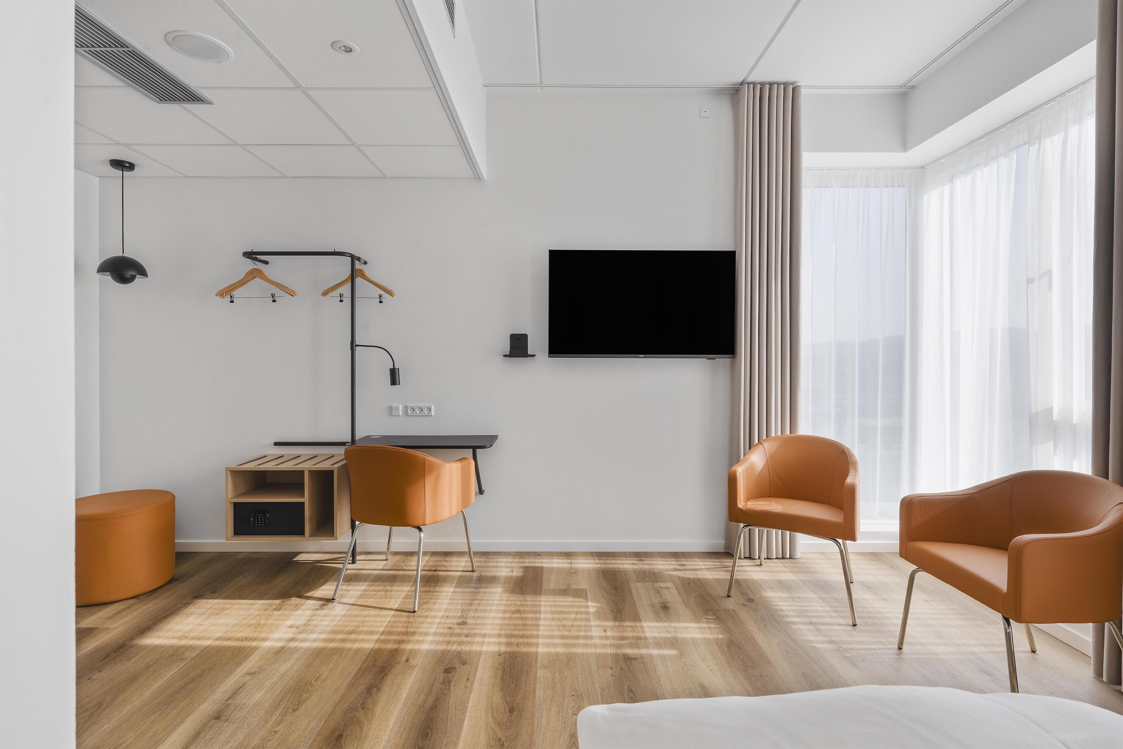 A modern hotel room with a wooden floor, a desk, two orange chairs, a TV, and a large window with curtains.