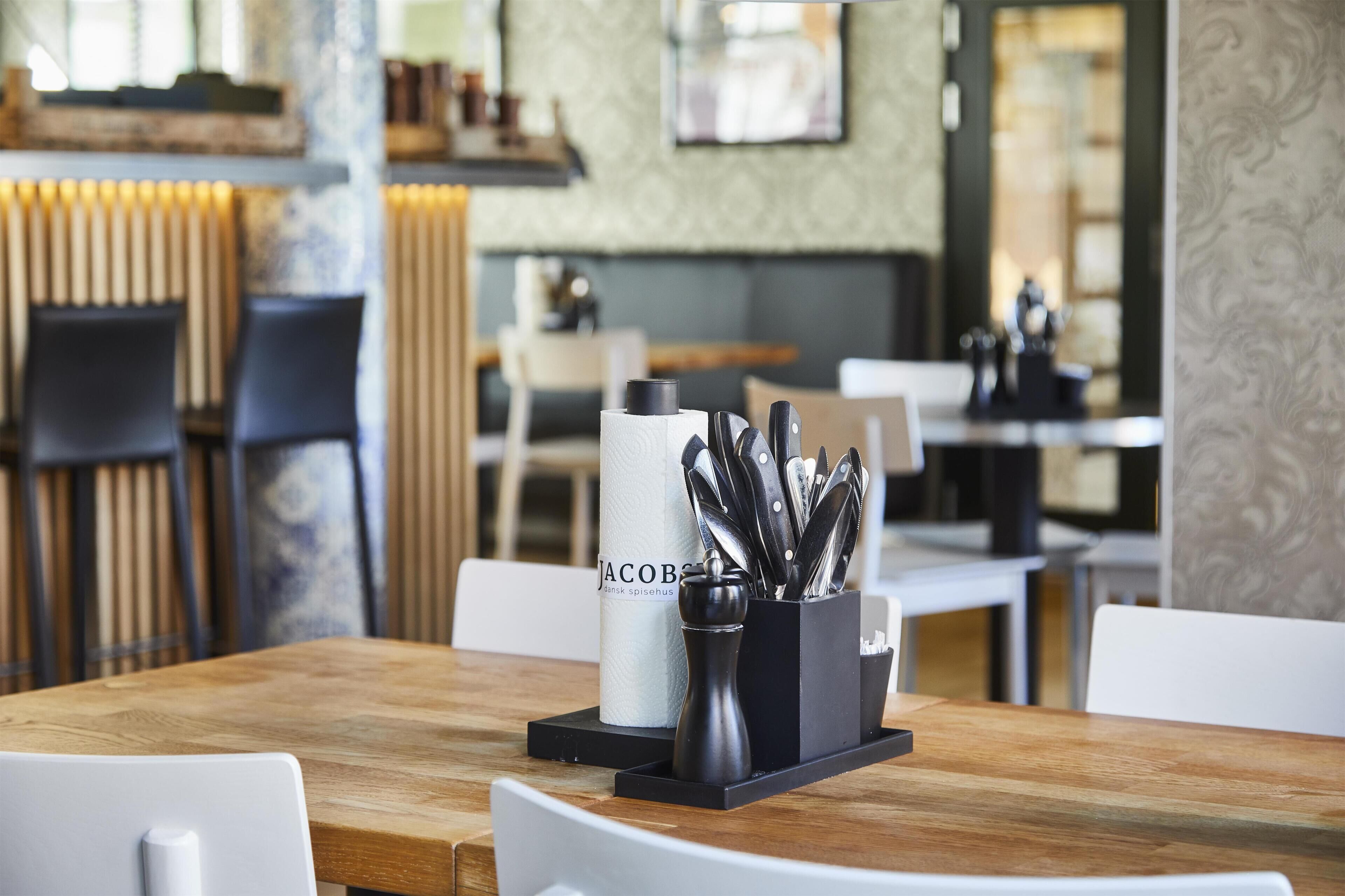 Ein Restaurantinnenraum mit einem Holztisch, einem schwarzen Besteckhalter und einem Papierhandtuchspender mit der Aufschrift 'Jacob's'.