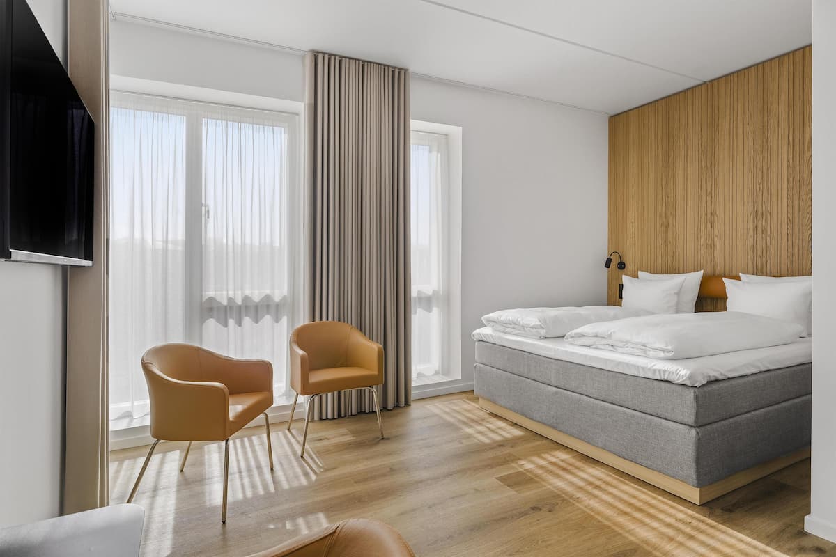 A modern hotel room with a bed, a chair, a TV, and large windows.