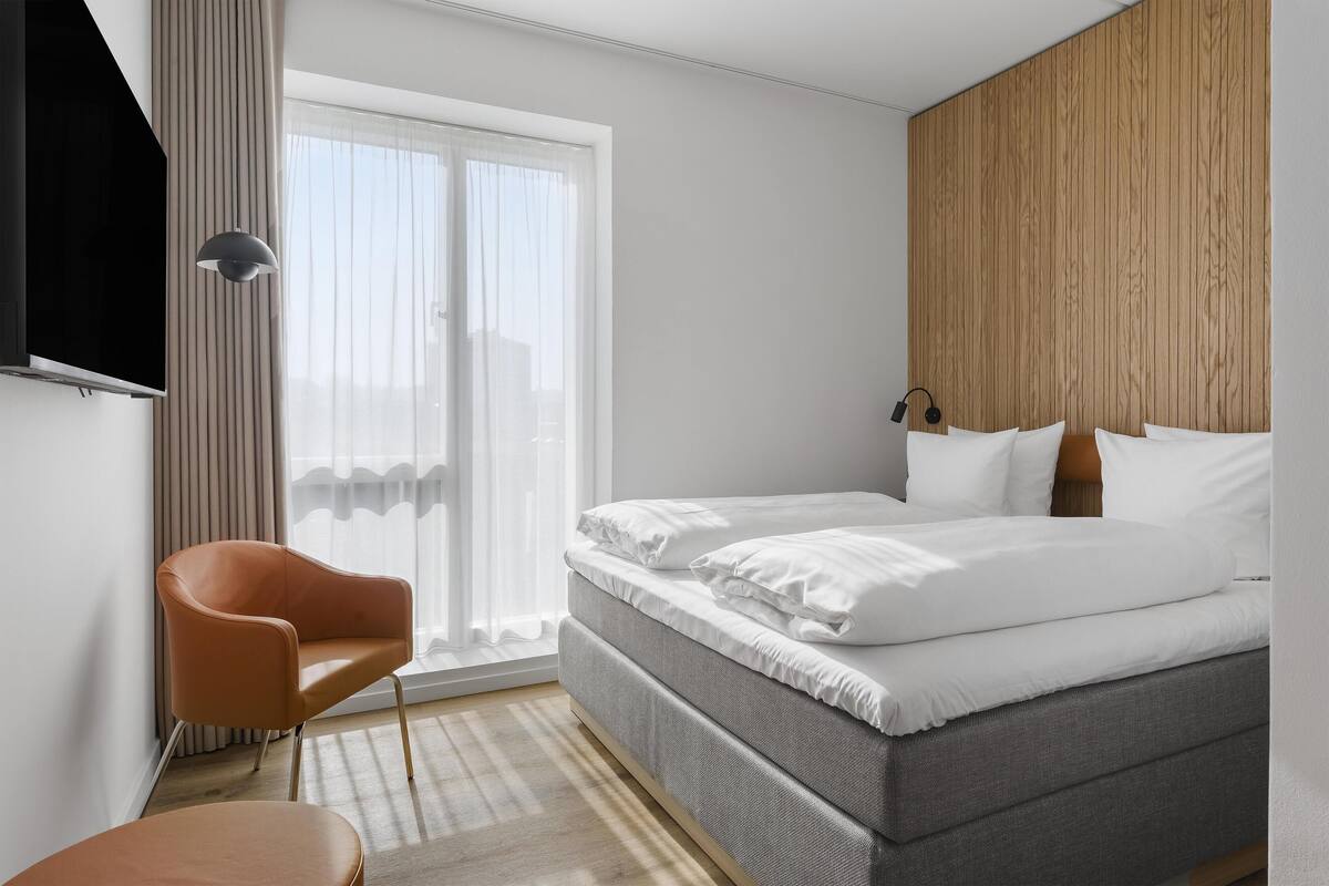 A modern hotel room with a bed, a chair, a TV, and a wooden accent wall.