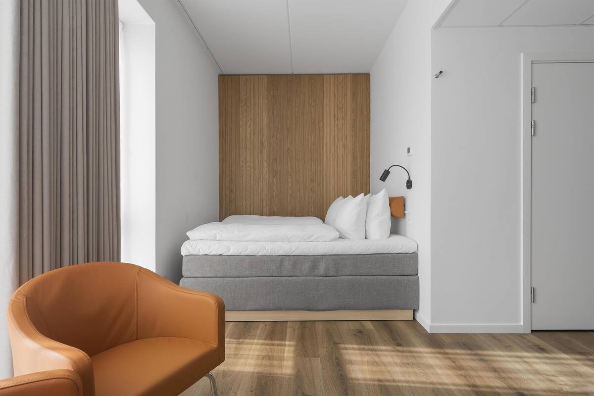 A modern hotel room with a bed, a chair, a wooden panel wall, and a door.