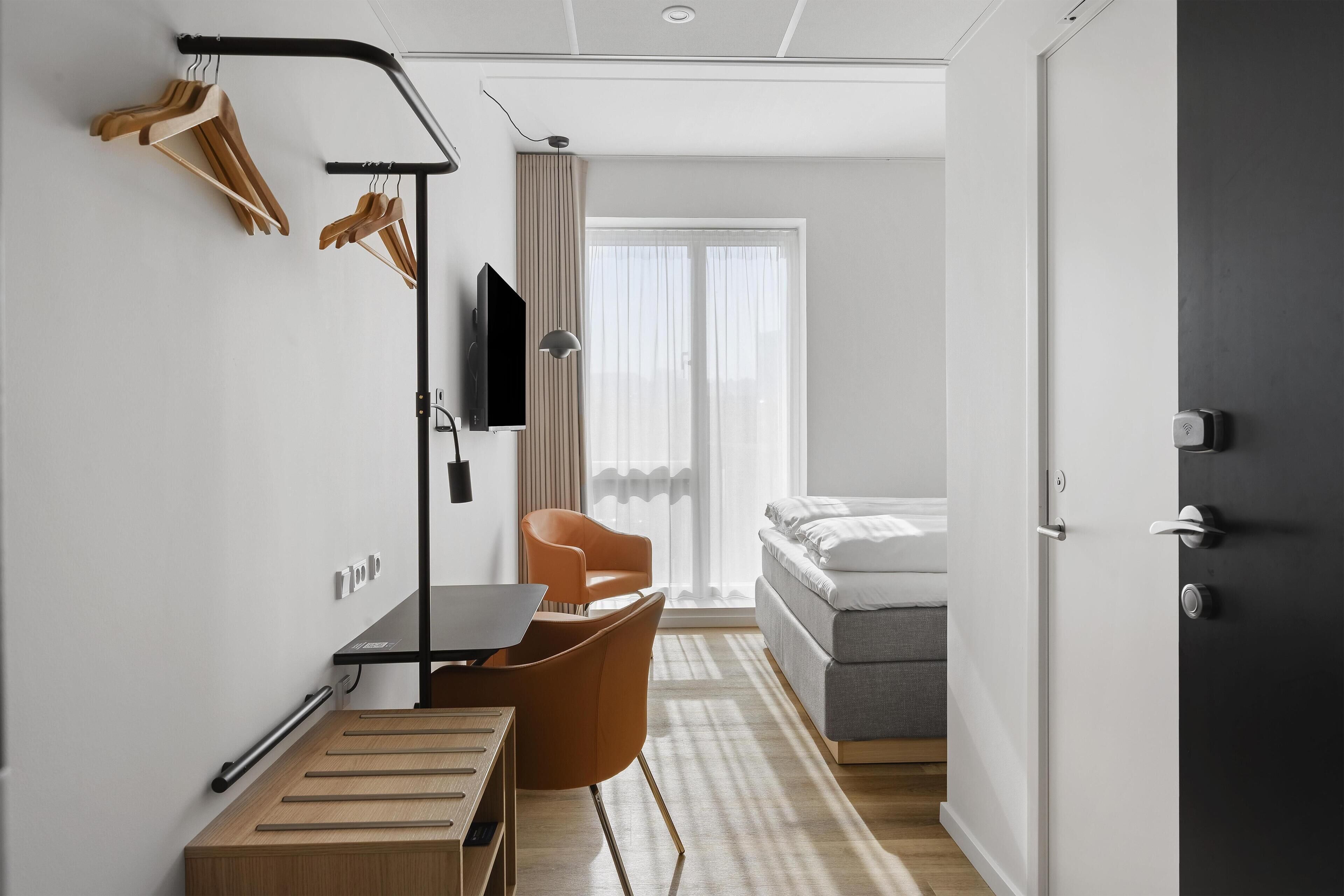 A modern hotel room with a desk, chair, bed, and a view of the outdoors.