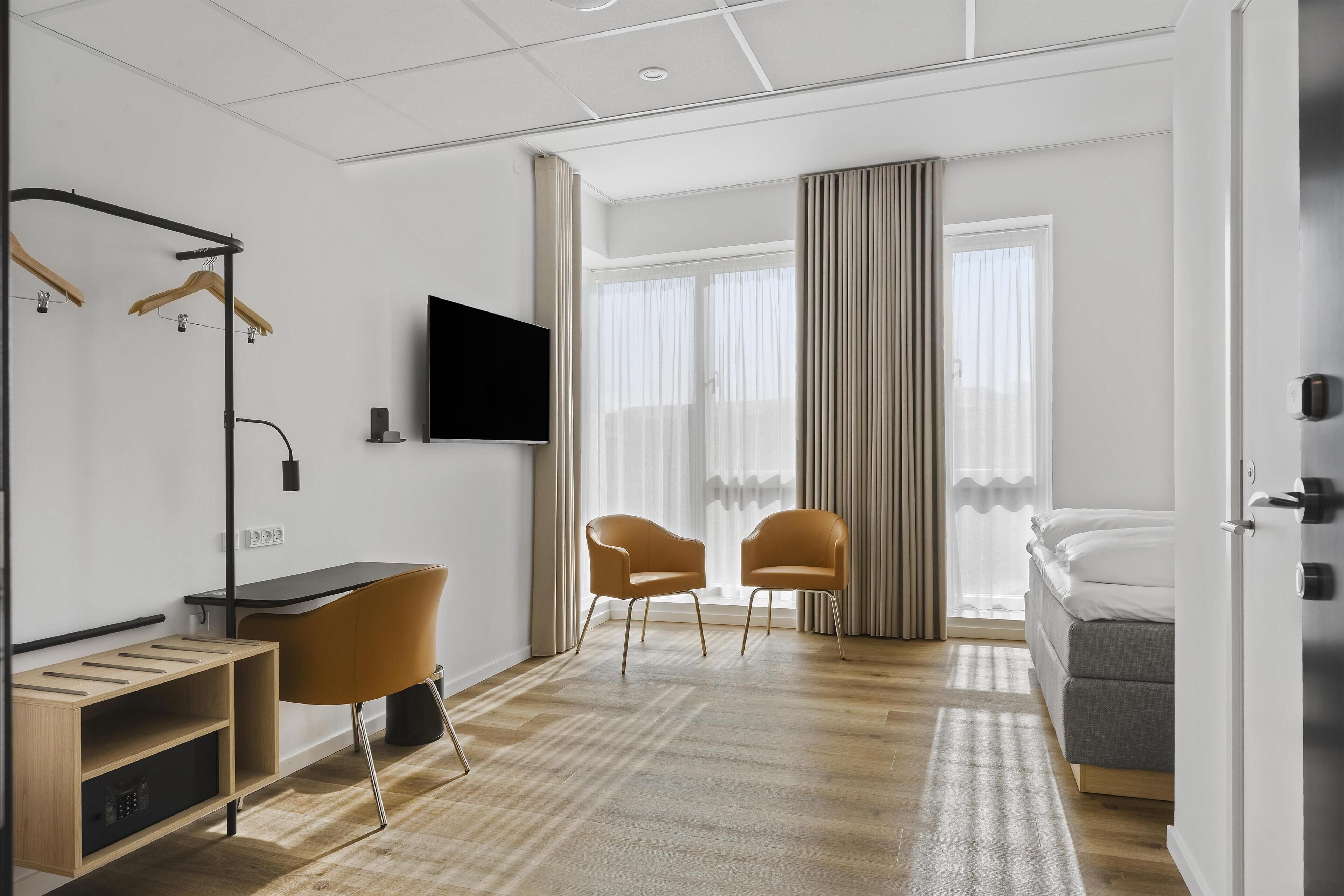 Una camera d'albergo moderna con una scrivania, due poltrone, un letto, una televisione e una finestra con tende.