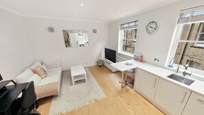 Een moderne, goed verlichte kamer met een witte bank, een klein wit tafeltje, een televisie, een wit bureau en een raam met uitzicht op een bakstenen gebouw.