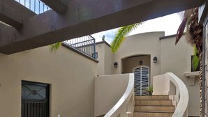 Un escalier menant à une porte surmontée d’un portail en métal, le tout entouré d’une terrasse dallée de pierres et d’un espace couvert doté d’un toit incurvé.